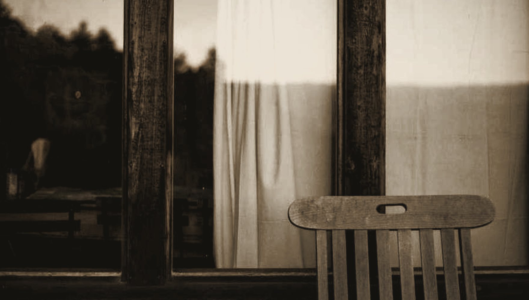 A spare, sunlit interior — a single chair beside a tall window, linen curtain breathing.